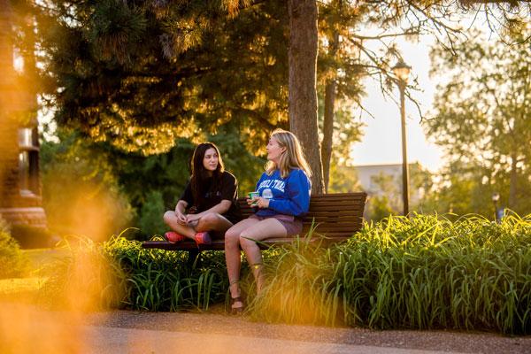 Two students seated on a bench talking, with trees and plants around them